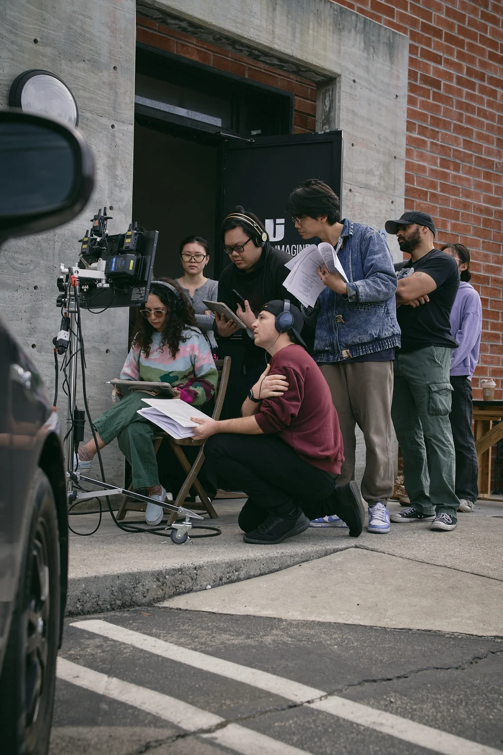 Christian Gonzales kneeling with a script beside a film crew gathered around a monitor outdoors